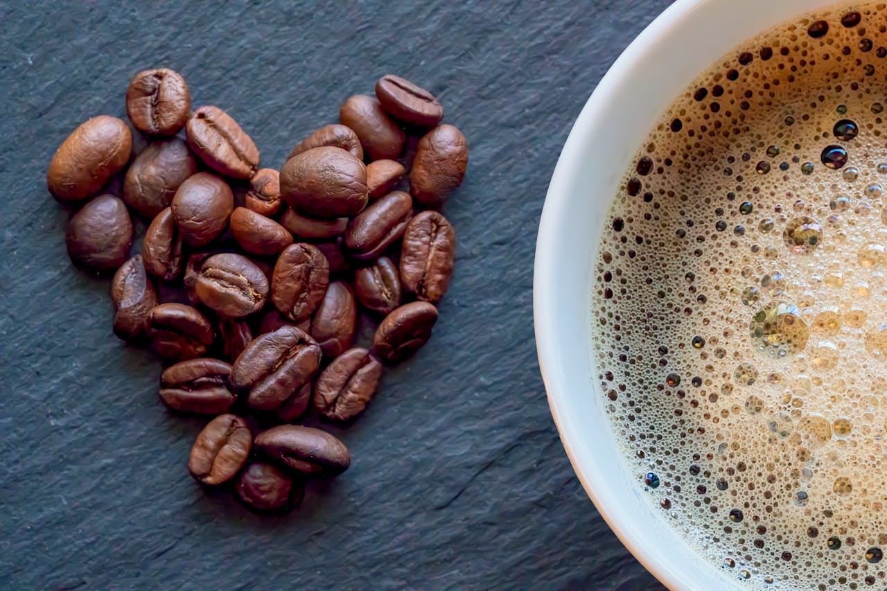 services-03 Close-up of heart-shaped coffee beans next to a cup of frothy coffee on a slate surface.